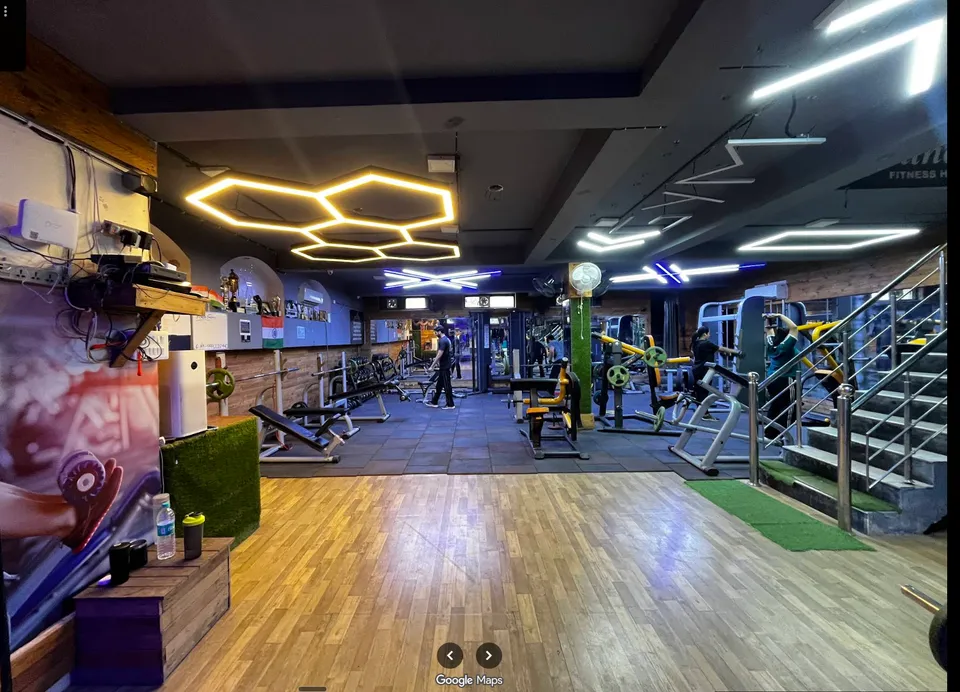 A member walking past cable and weight machines on the gym floor at Fitness Factor, with the staircase and gym signage in the background.
